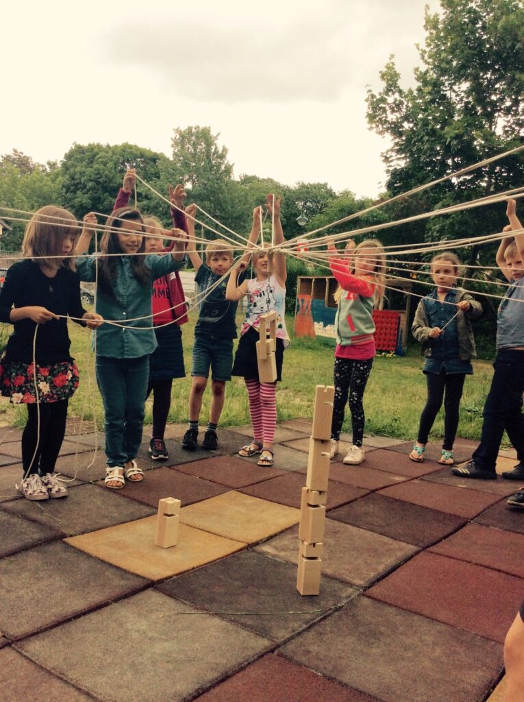 Kinder einer Schulklasse lösen gemeinsam ein Teamspiel mit Seilen im Hof des Yellow